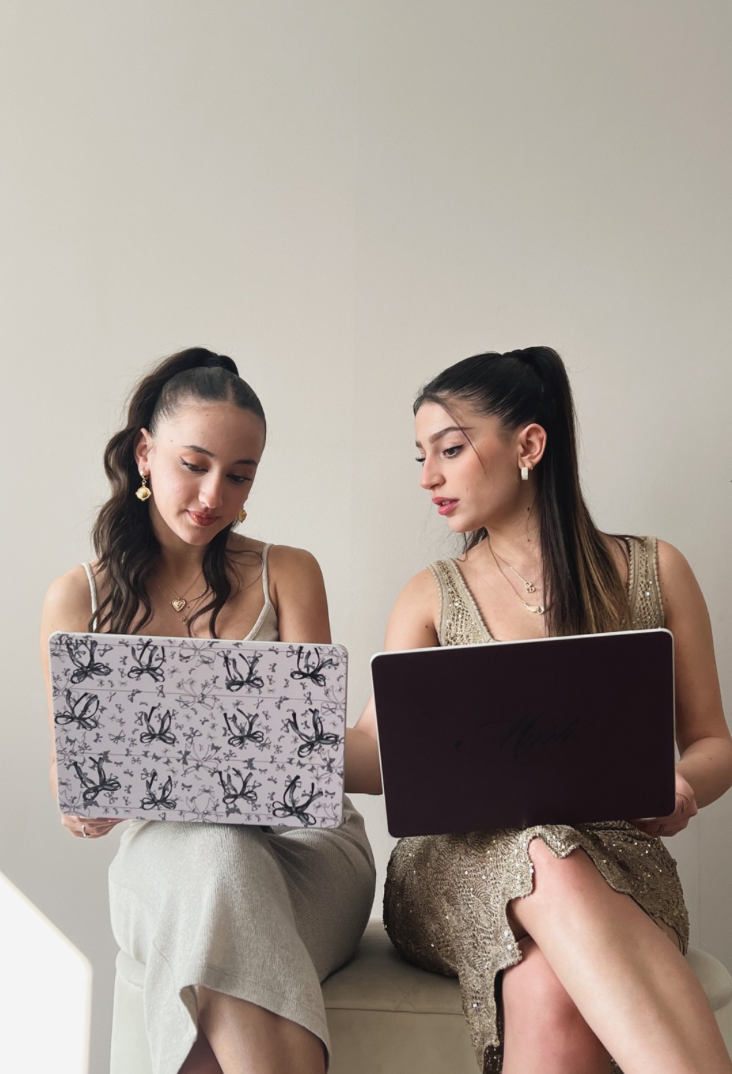 "Icon featuring two girls in formal dresses sitting together, each holding stylishly designed MacBook cases against a beige background, highlighting the elegant laptop accessories."